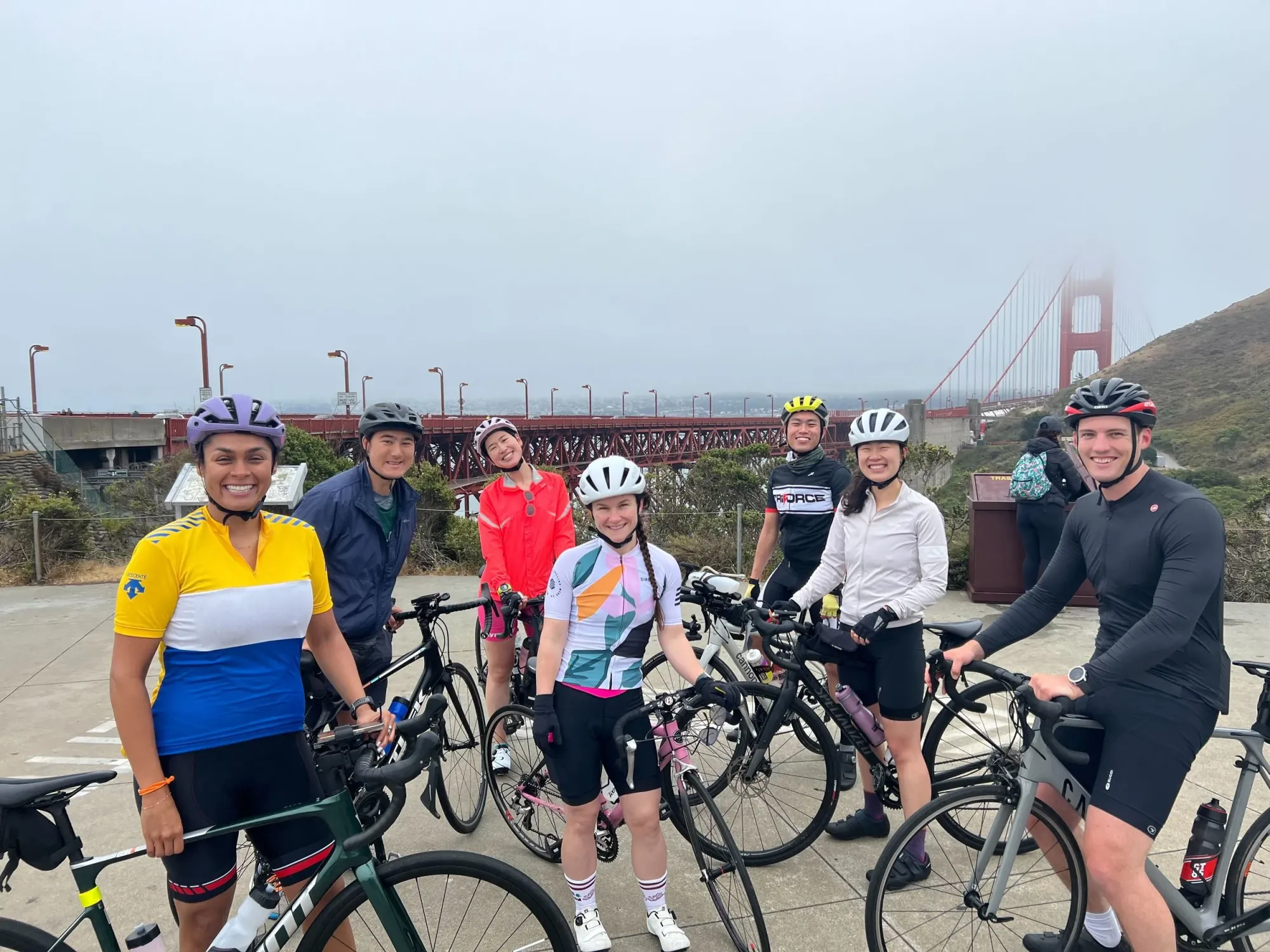 Eight SF Tri Club cyclists standing with their road bikes at a viewpoint in front of a fog-shrouded Golden Gate Bridge.