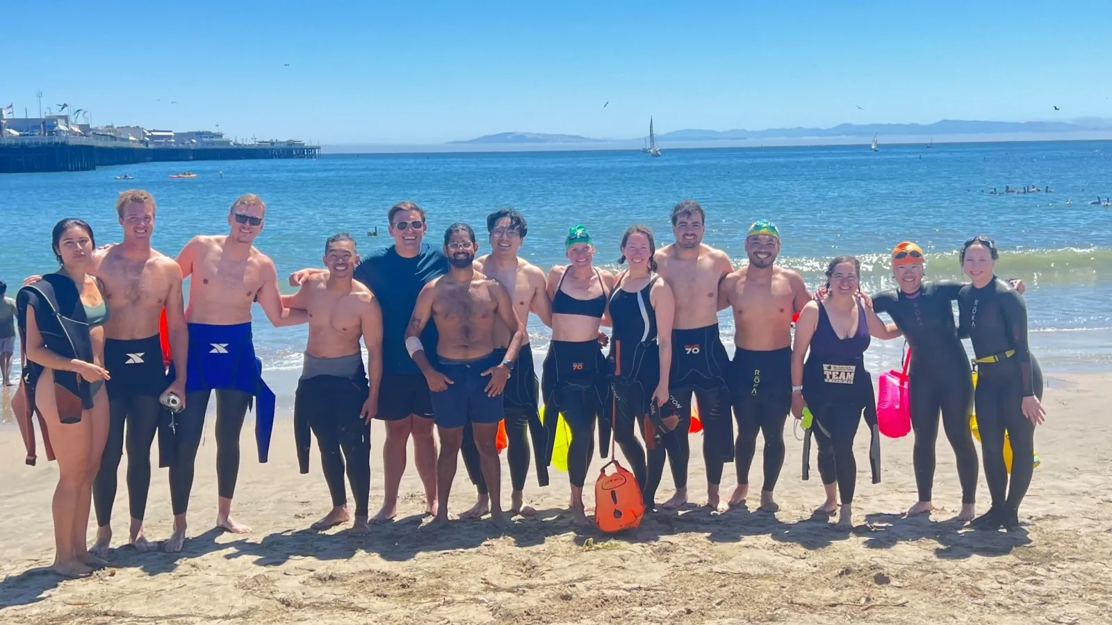 A group of SF Tri Club athletes in wetsuits on the beach after an open water swim at Santa Cruz, with the wharf behind them.